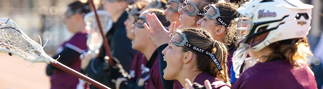 Women's lacrosse players cheer on their teammates from the slidelines.