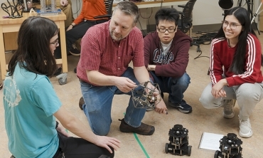 A professor and students crouch down to examine a pair of AI-powered robots.